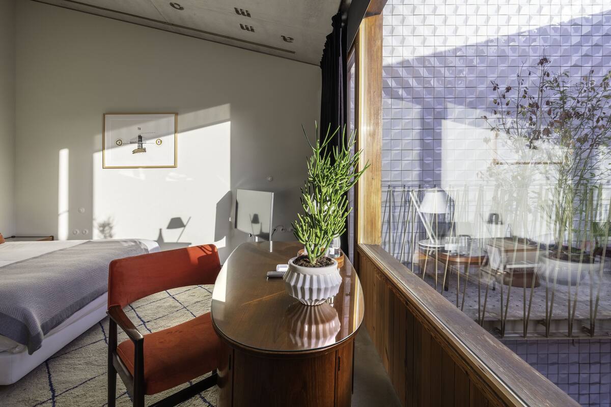 Un dormitorio con cama, una mesita de noche de madera con una planta en maceta, una silla roja y una ventana con vistas a un balcón amoblado.