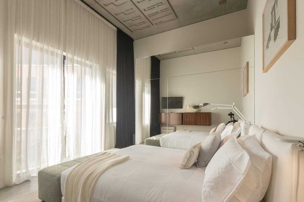 Un dormitorio moderno con una cama grande, una mesita de noche, un espejo y una ventana con cortinas translúcidas.