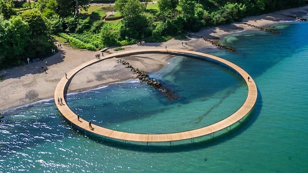 An aerial view of a circular wooden deck extending into a clear blue body of water, surrounded by a sandy beach and greenery.