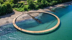An aerial view of a circular wooden deck extending into a clear blue body of water, surrounded by a sandy beach and greenery. - Hørhavegården (Aarhus)