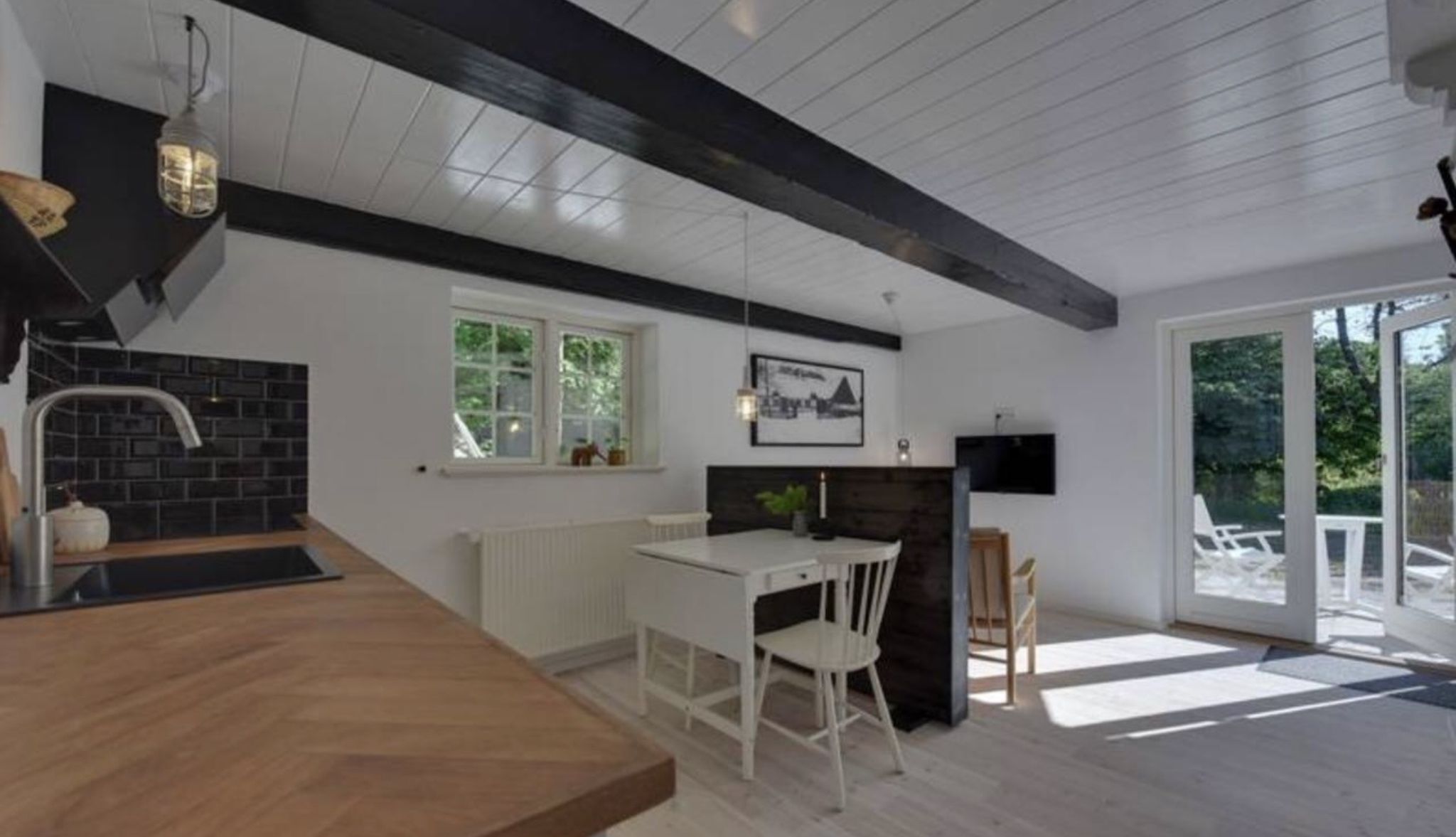A modern kitchen with a wooden countertop, white cabinetry, and a black backsplash. There is a dining area with a white table and chairs, and a large window providing natural light.