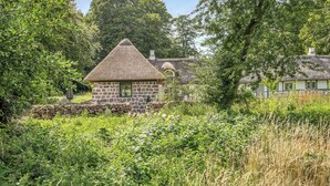 A stone cottage with a thatched roof, surrounded by lush greenery and trees.