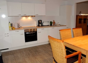 A modern kitchen with white cabinets, a built-in oven, a coffee maker, and a dining area with wicker chairs and a wooden table. - Vacation apartment Landliebe boben - Holiday apartments Landliebe, 15187 (Leer (Ostfriesland))