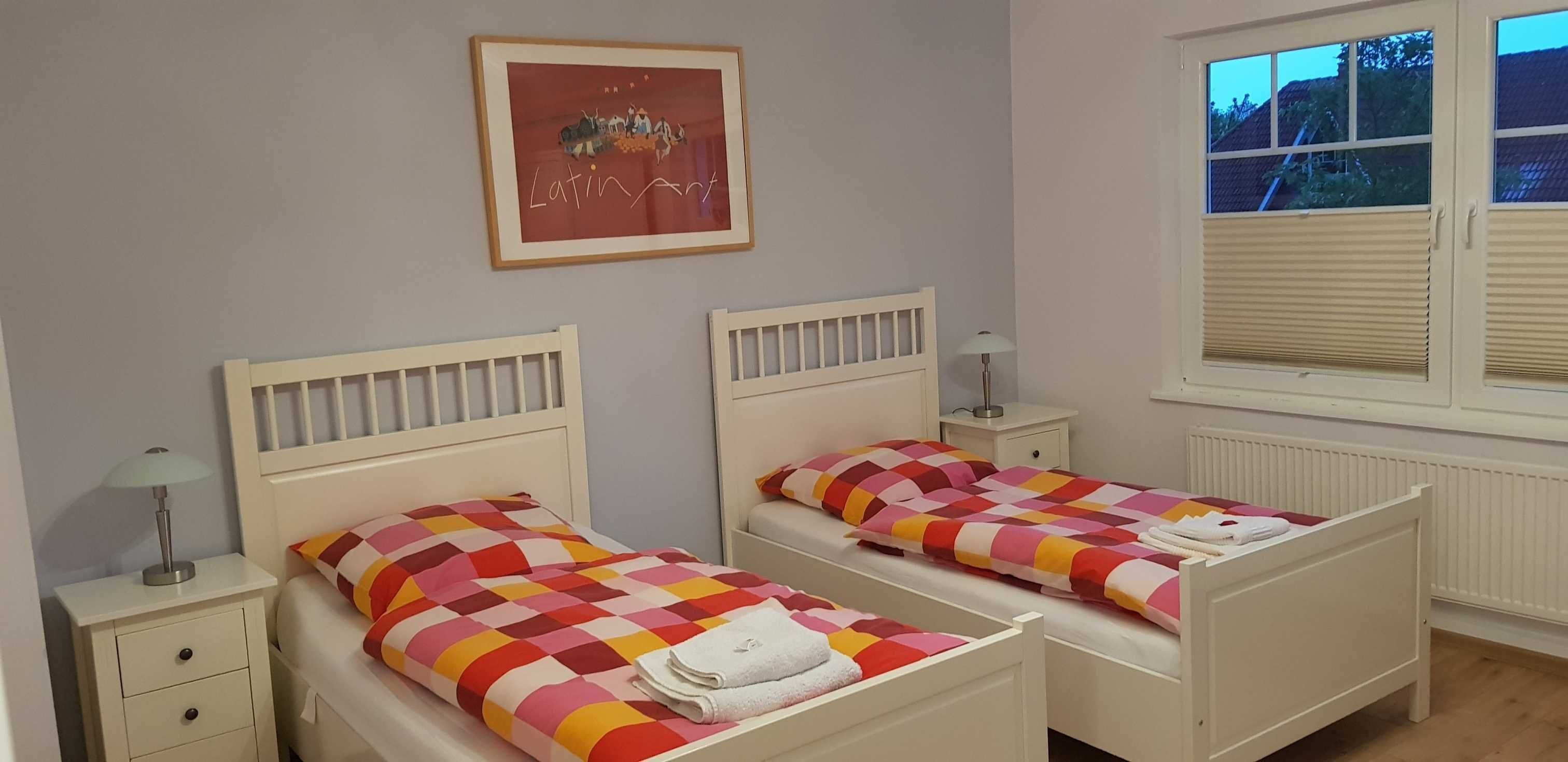 A room with two single beds, a nightstand with a lamp, a framed picture on the wall, and a window with blinds.