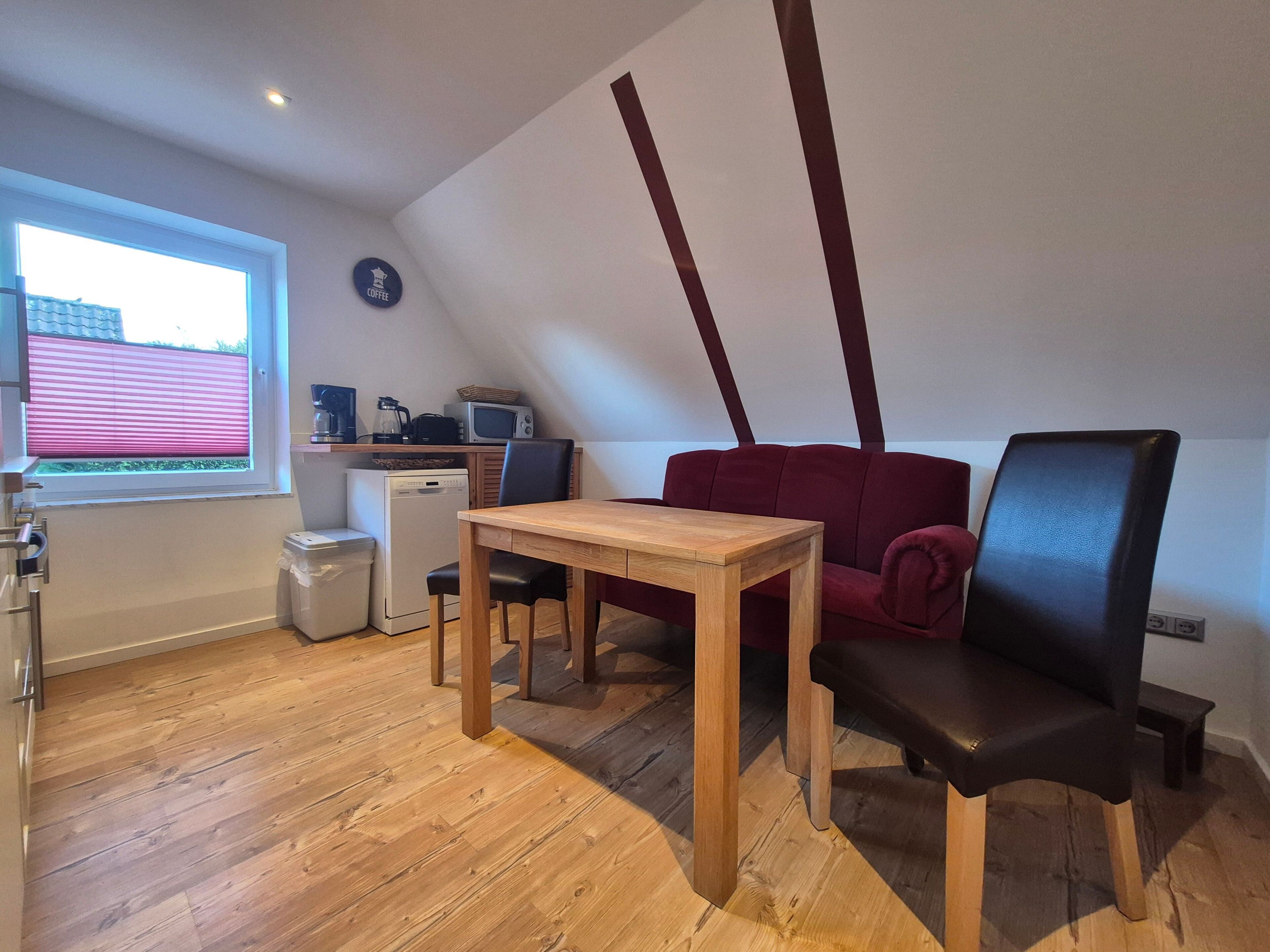 A compact living space with a dining table, a red sofa, a black chair, a small kitchenette, and a window with blinds.