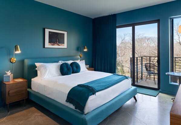 A modern bedroom with a large bed, two bedside lamps, a framed picture, and a balcony with a view.