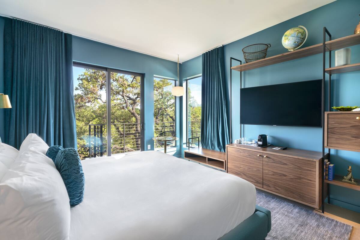 Ein modernes Hotelzimmer mit einem großen Bett, einem Fernseher, einem Holzschrank und einem Balkon mit Blick auf Bäume.