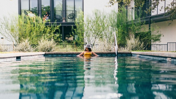 A swimming pool with a person in the water, surrounded by greenery and a building in the background. - The Ruby Hotel (Round Rock)