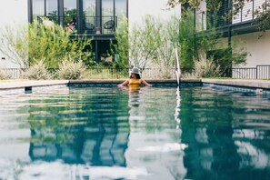 A swimming pool with a person in the water, surrounded by greenery and a building in the background.