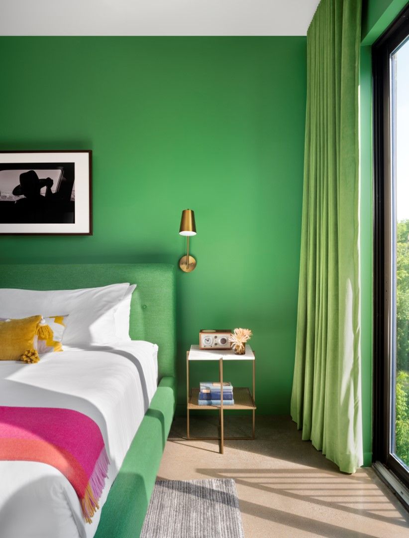 Une chambre avec un mur vert, un lit avec une literie blanche, une table de chevet avec une lampe et une fenêtre avec des rideaux.