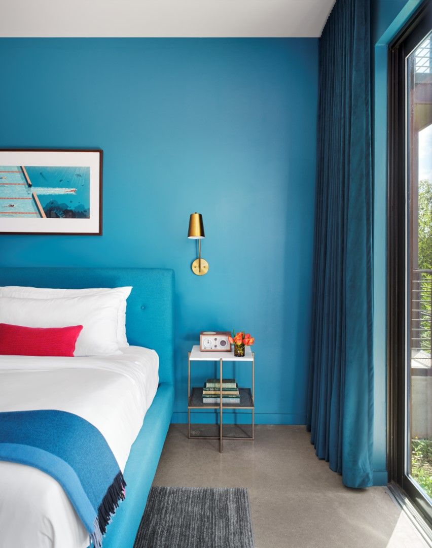 A bedroom with a bed, a bedside table with a radio, a framed picture on the wall, and a window with curtains.