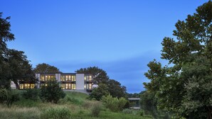 A modern two-story house with large windows, surrounded by lush greenery and a river. - The Ruby Hotel (Round Rock)