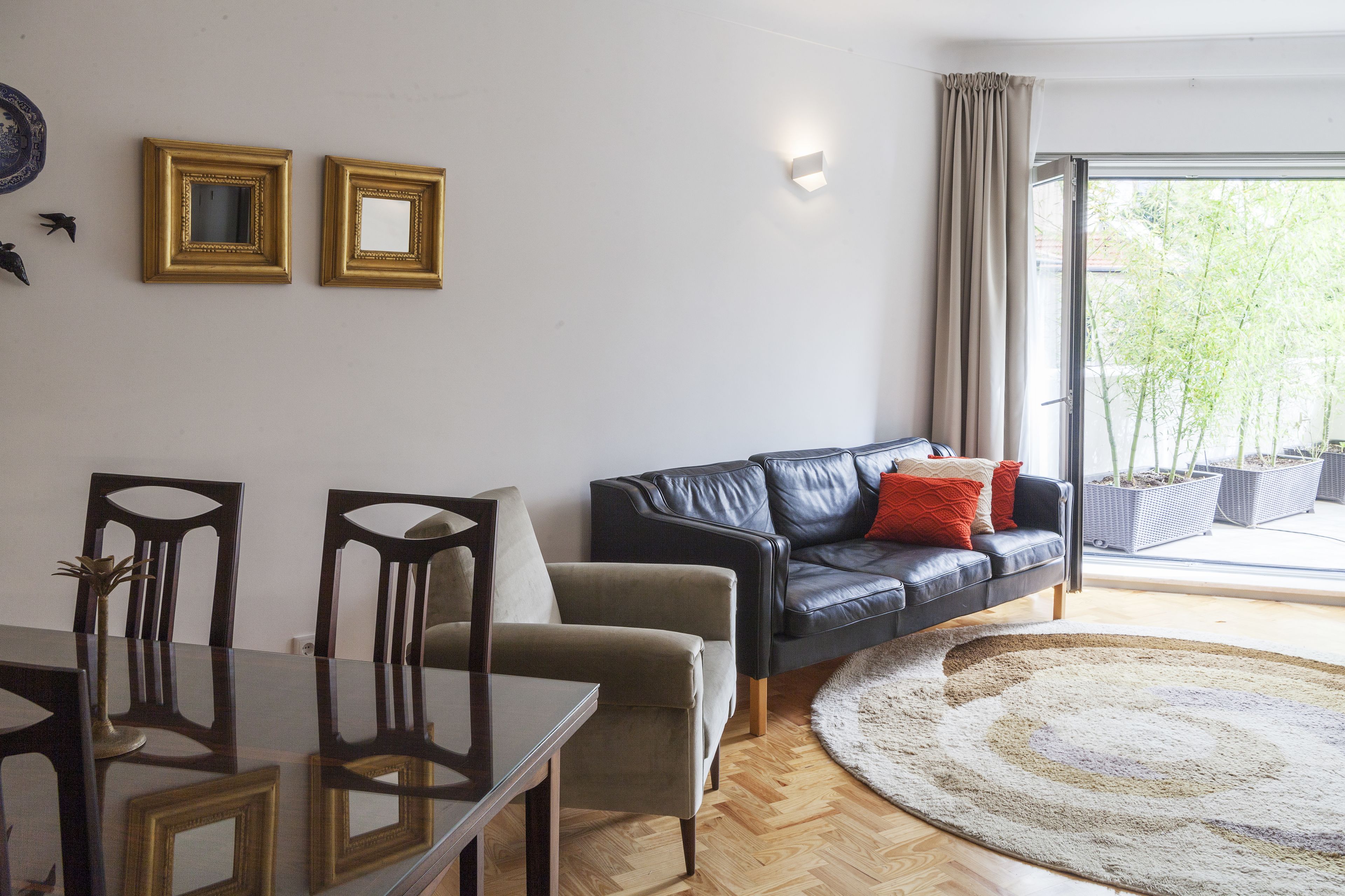 Ein Wohnzimmer mit Esstisch, zwei Stühlen, einem schwarzen Ledersofa mit roten Kissen und einem großen Fenster mit Blick auf Bäume.