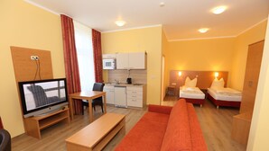 Una habitación de hotel compacta con sofá, televisor, pequeña mesa de comedor, kitchenette y dos camas.