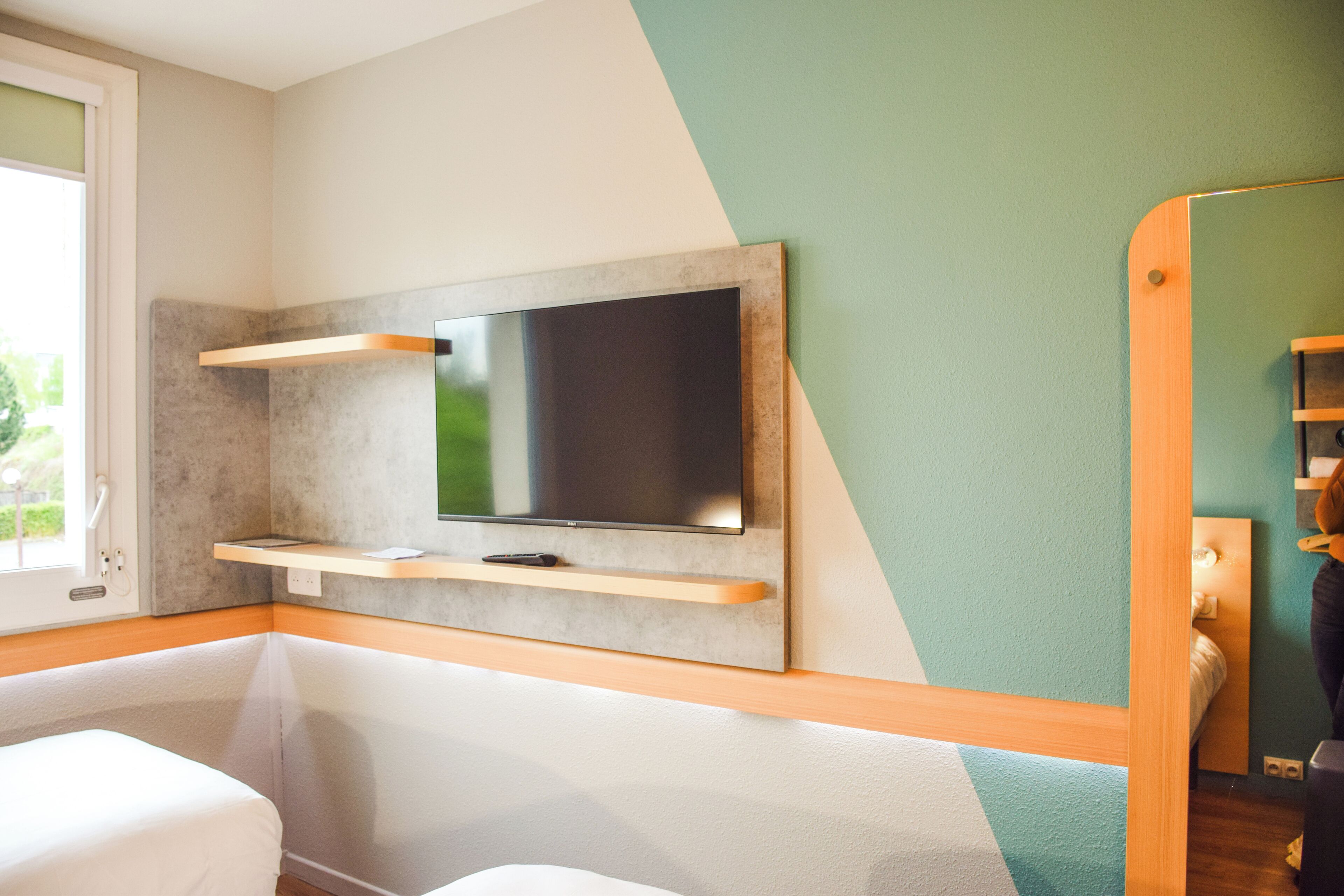 A modern hotel room with a flat-screen TV mounted on a wall, a wooden shelf below it, and a bed with a white headboard.