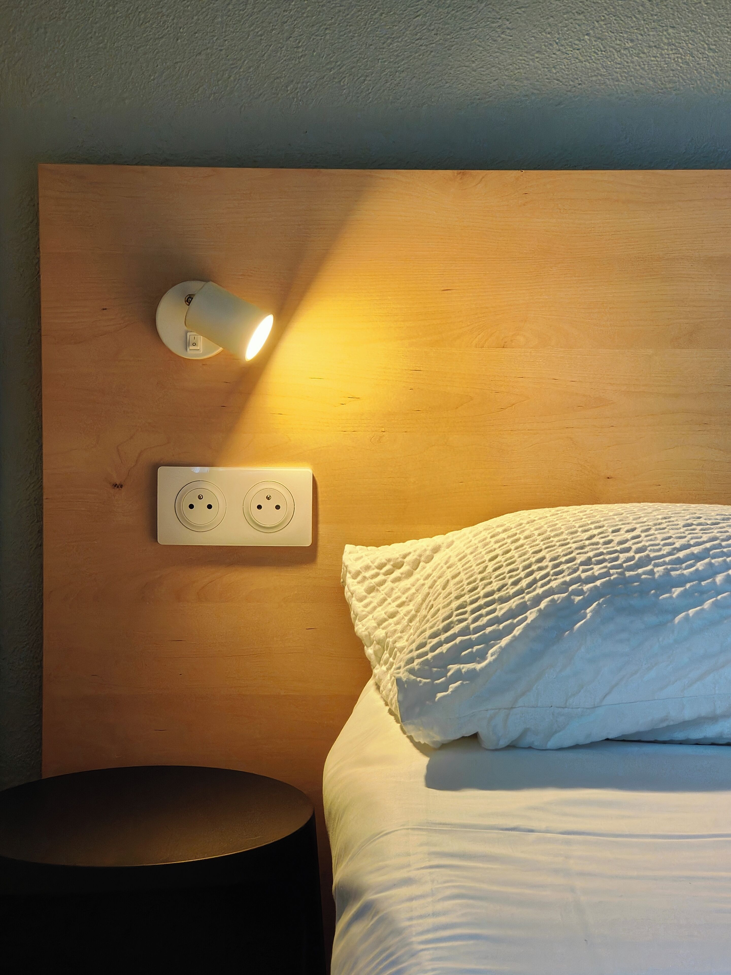 Una cama bien hecha con un almohadón blanco texturizado, un enchufe y una lámpara de pared.