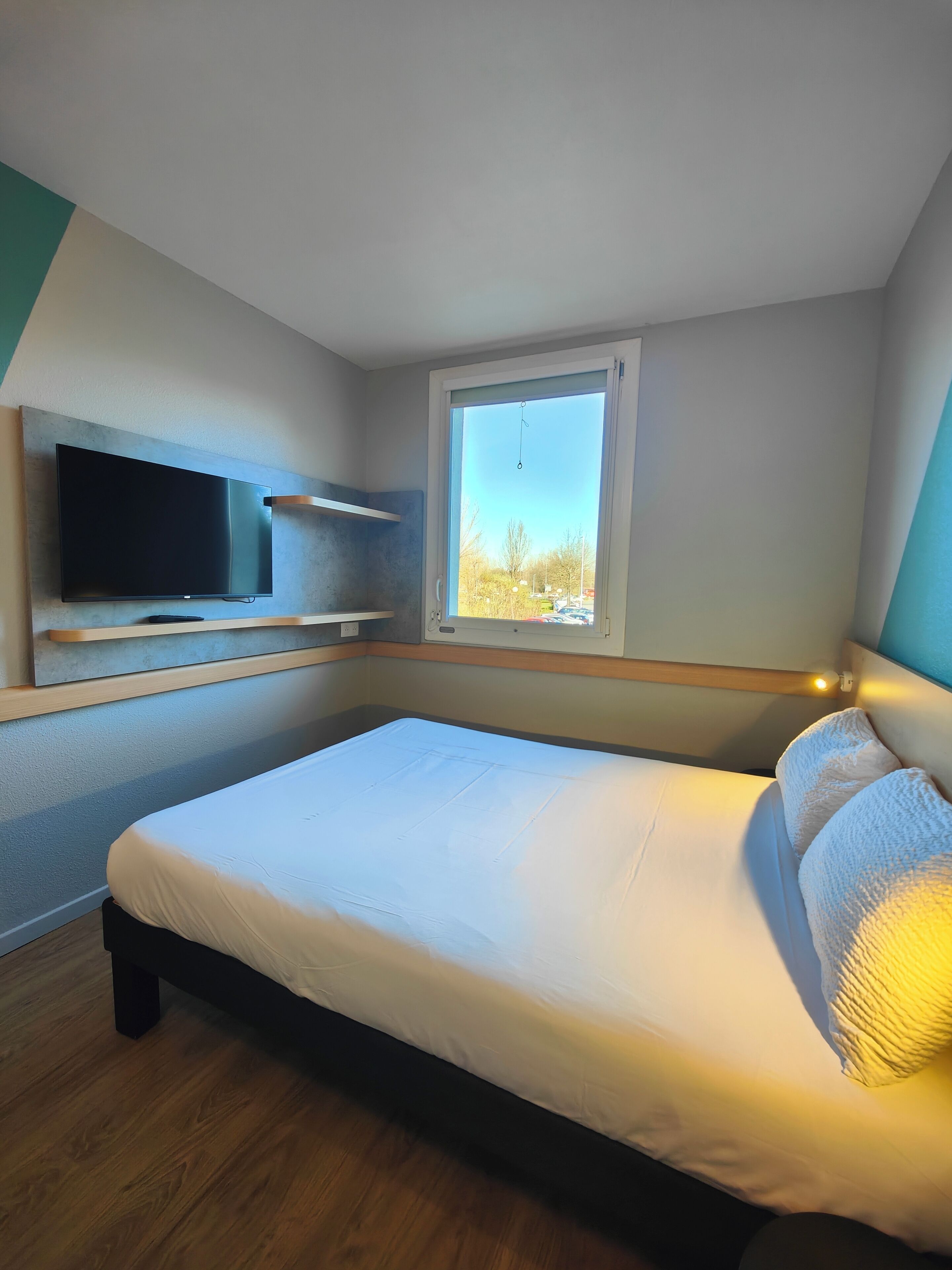 Une chambre moderne avec un lit, un téléviseur fixé au mur et une fenêtre donnant sur l’extérieur.