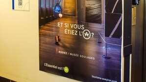 A hallway with a large advertisement for Aveyron, featuring a person in a blue outfit and a hat, a staircase, and a QR code on a sign. - Ibis Budget Rodez (Rodez)
