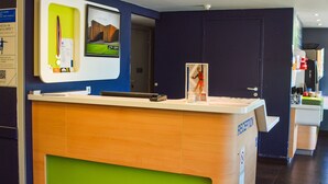 A reception desk with a green accent, a digital screen, and a door leading to a hallway. - Ibis Budget Rodez (Rodez)