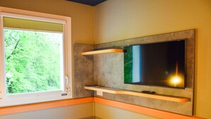 A room with a bed, a window with a view of greenery, a flat-screen TV mounted on a wall, and a small table. - Ibis Budget Rodez (Rodez)