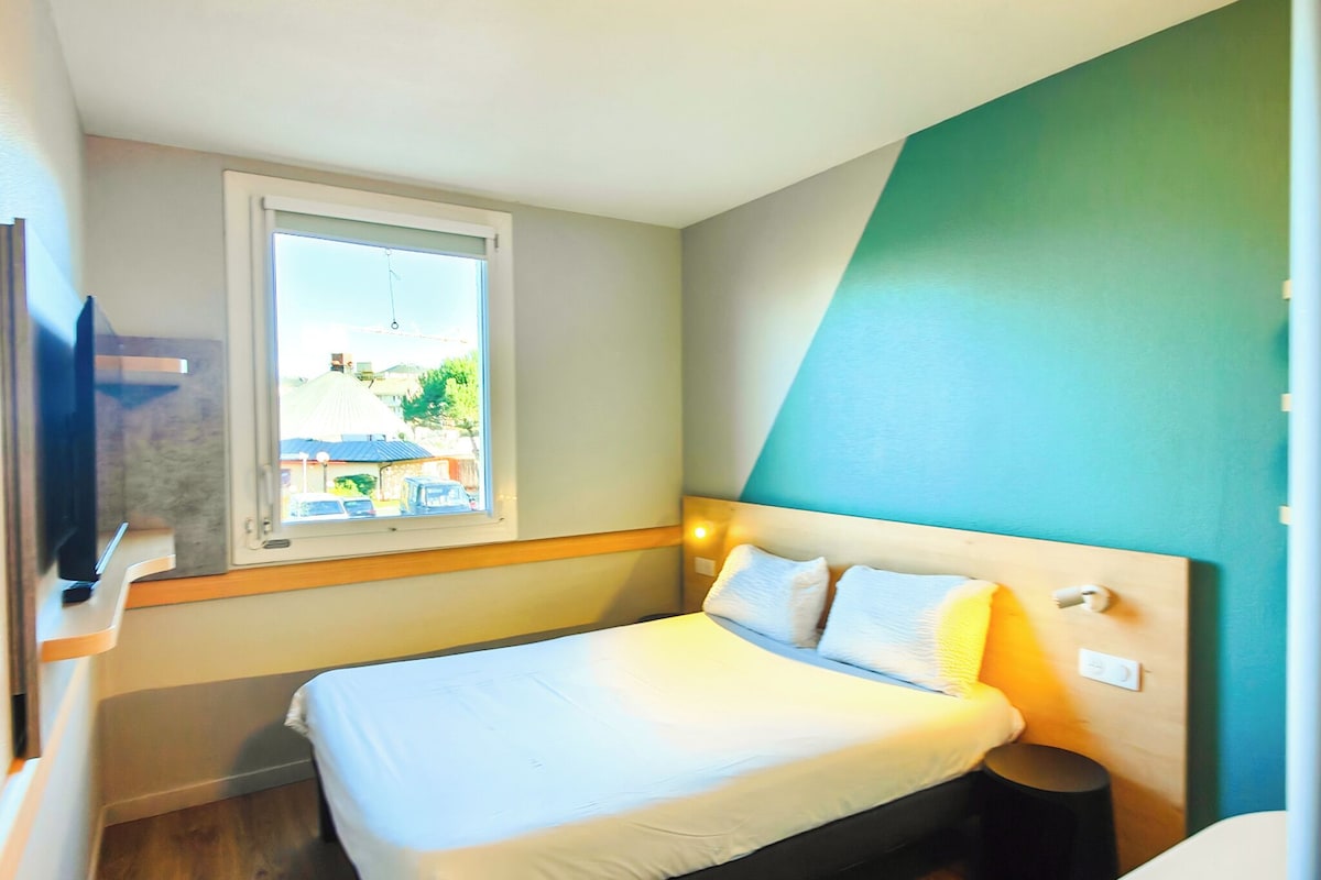 Un dormitorio con cama, ventana, mesita de noche y vistas al exterior.