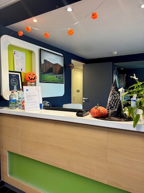 A reception desk with Halloween decorations, a framed certificate, and a digital screen displaying a building image. - Ibis Budget Rodez (Rodez)