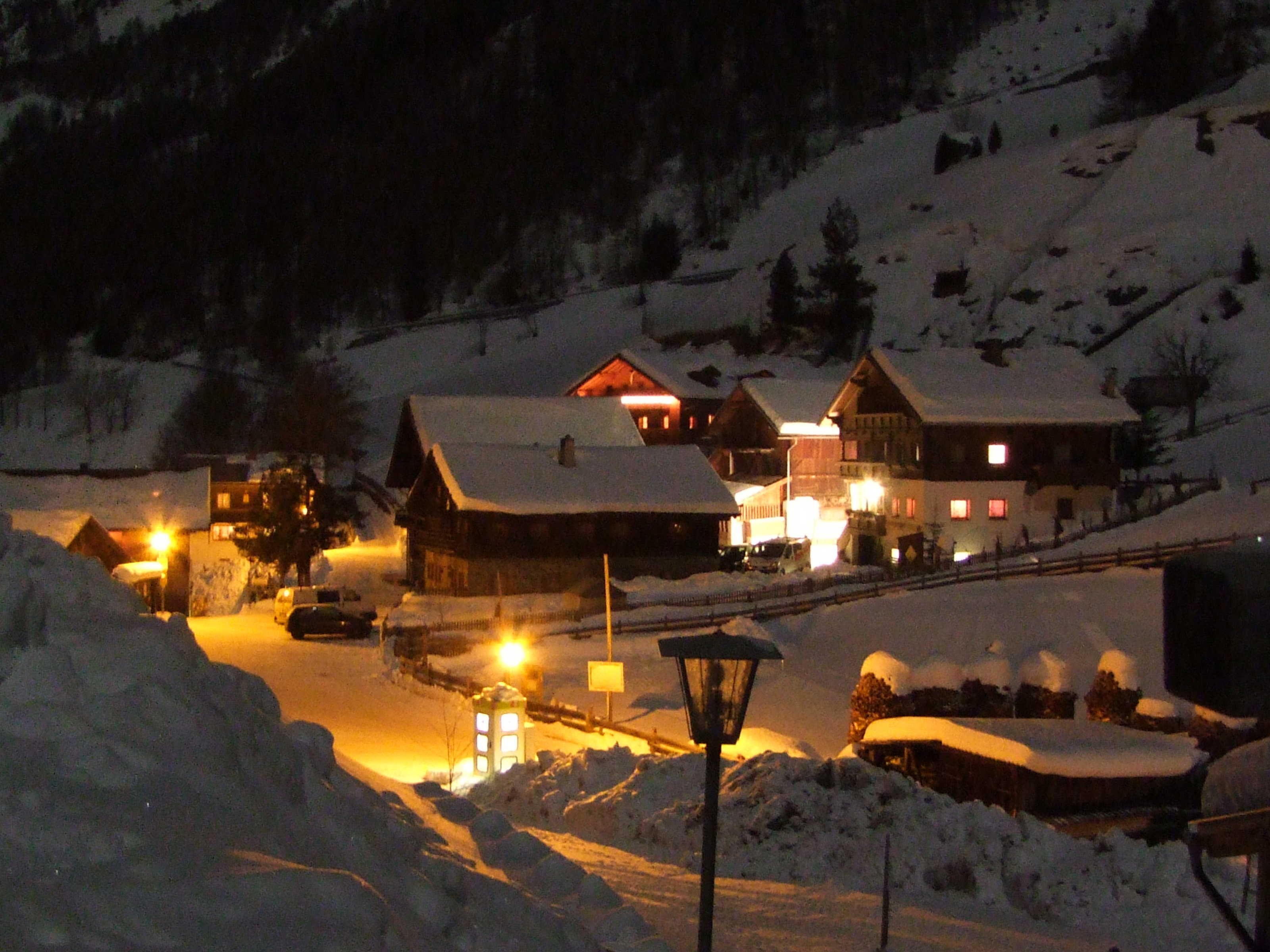 Un village enneigé avec des maisons traditionnelles en bois, une rue bordée de voitures et, au loin, une montagne recouverte de neige.