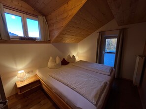 Un dormitorio con techo de madera, una cama con ropa de cama blanca, una mesita de noche con lámpara y una ventana con cortinas.