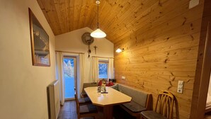 Una habitació acollidora amb sostre de fusta, taula de dinar amb cadires, una banca, finestra amb cortines i un llum penjant de la paret.