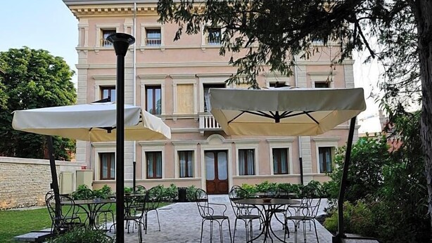 Un patio exterior con mesas y sillas, un edificio con balcones al fondo y un árbol en primer plano.