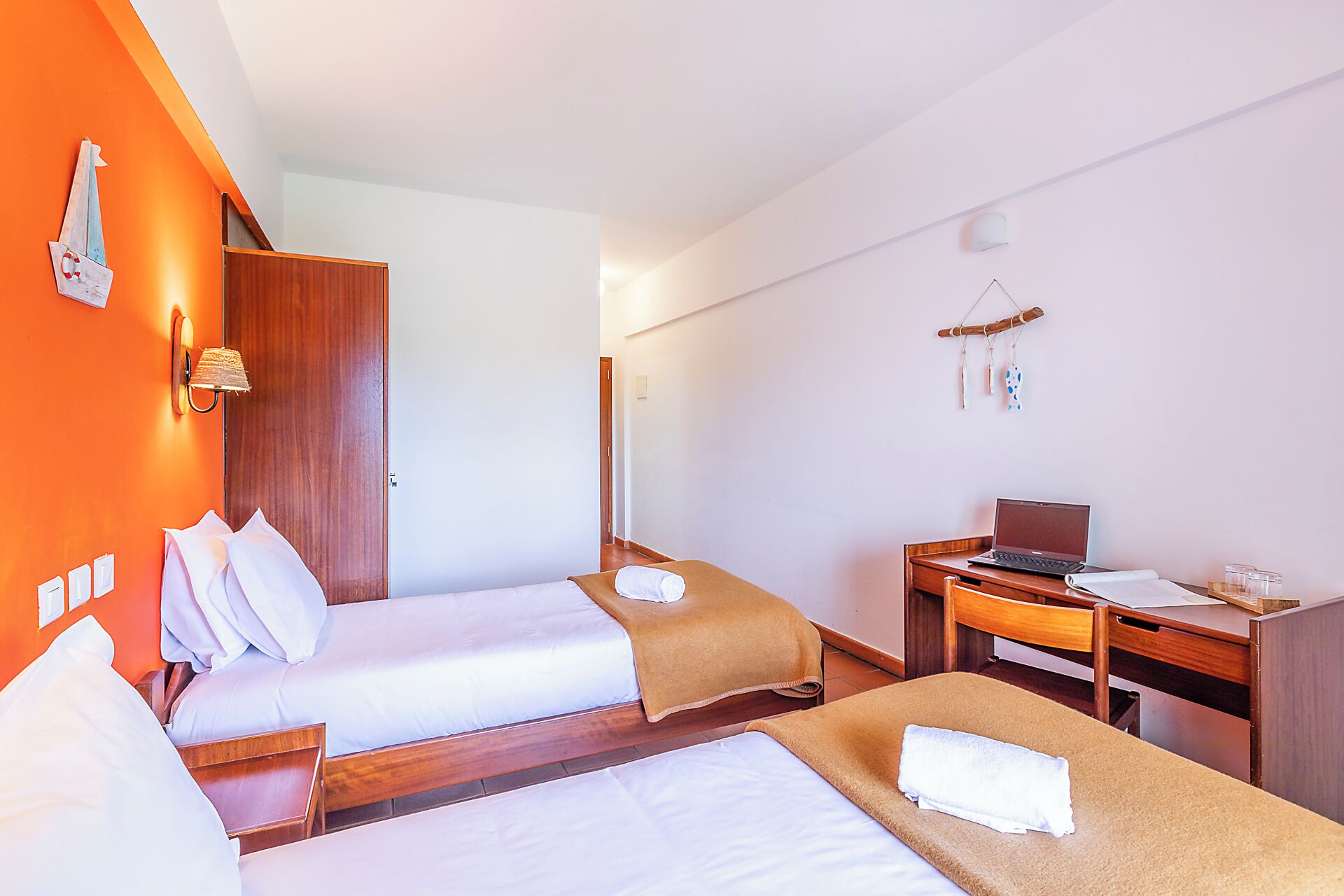 A hotel room with two beds, a desk, a laptop, and a wall-mounted lamp.
