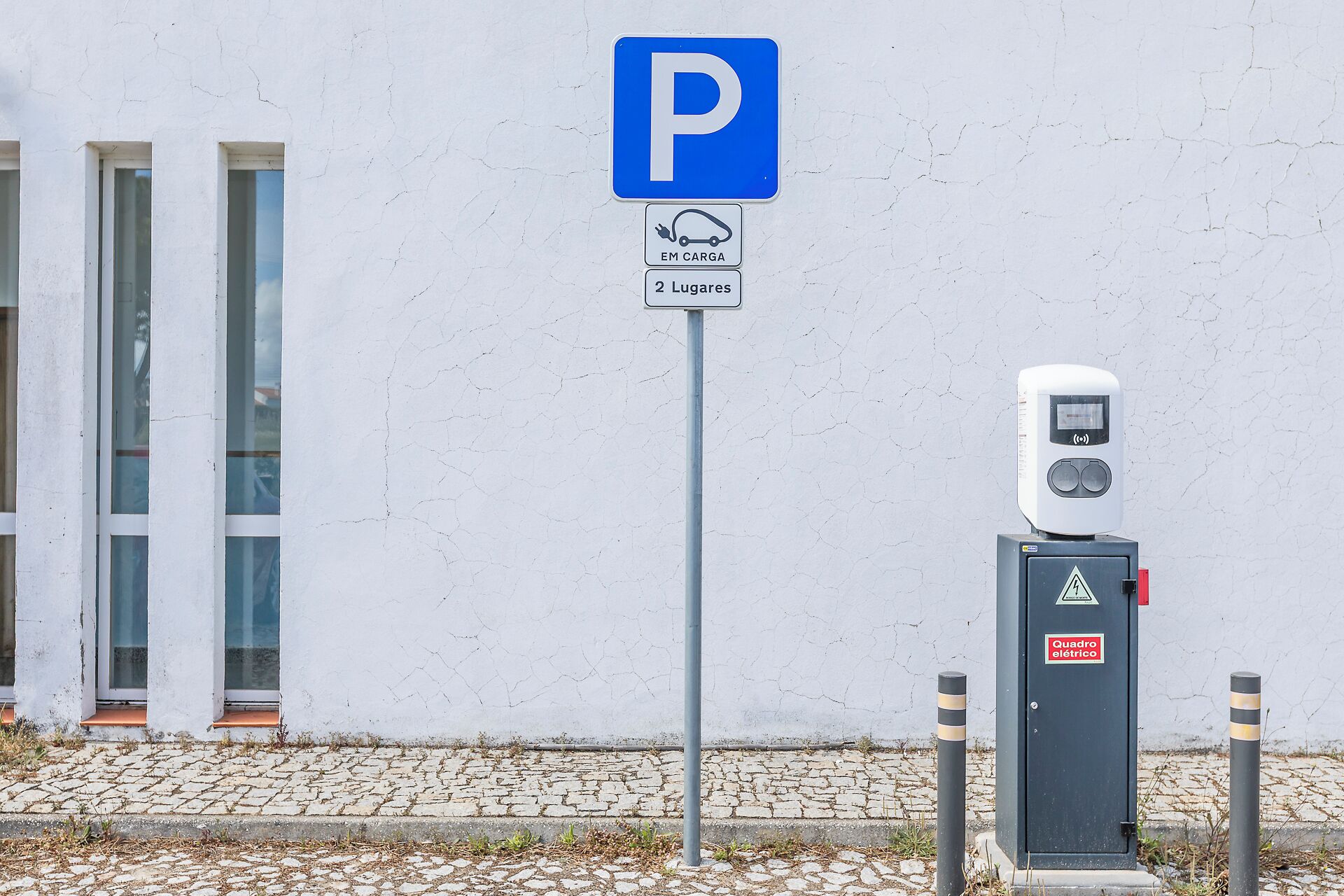 Segnale di parcheggio con simbolo di ricarica veicoli elettrici, una stazione di ricarica e un edificio con ampie vetrate.