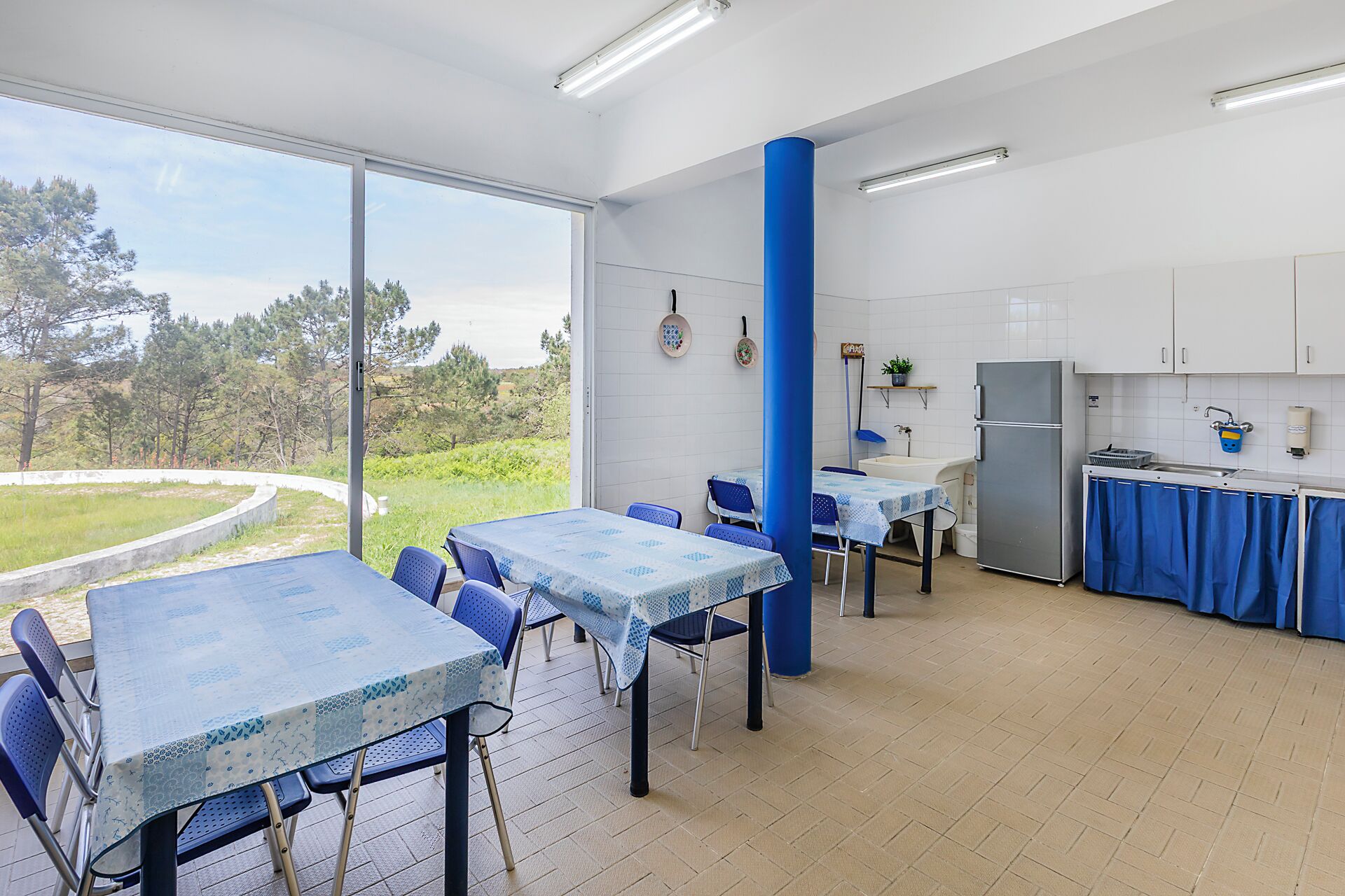 Zona pranzo con tavoli e sedie, cucina con frigorifero e vista sul verde.