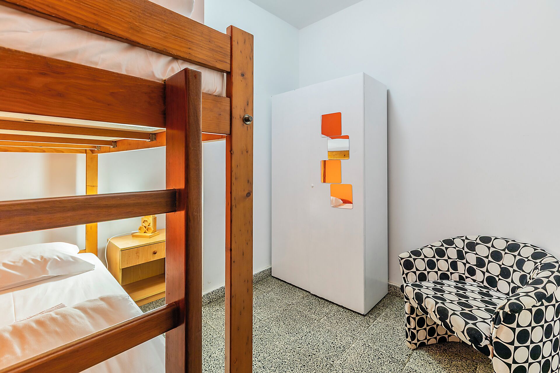 Quarto com cama de beliche, guarda-roupa branco e um banquinho estofado com padrão.