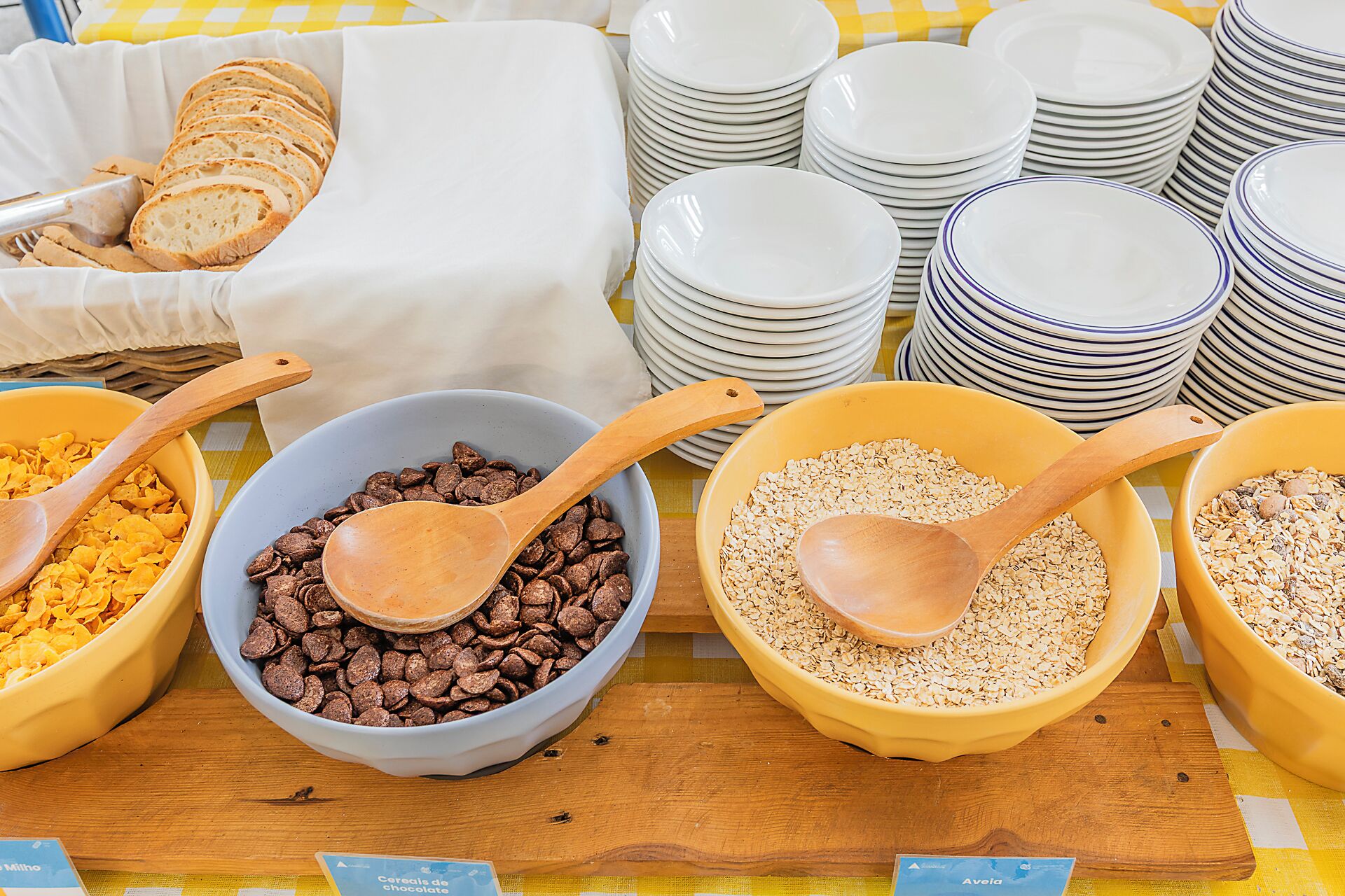 A buffet setup with bowls of cereal, chocolate chips, and oats, and stacks of white plates and bowls.