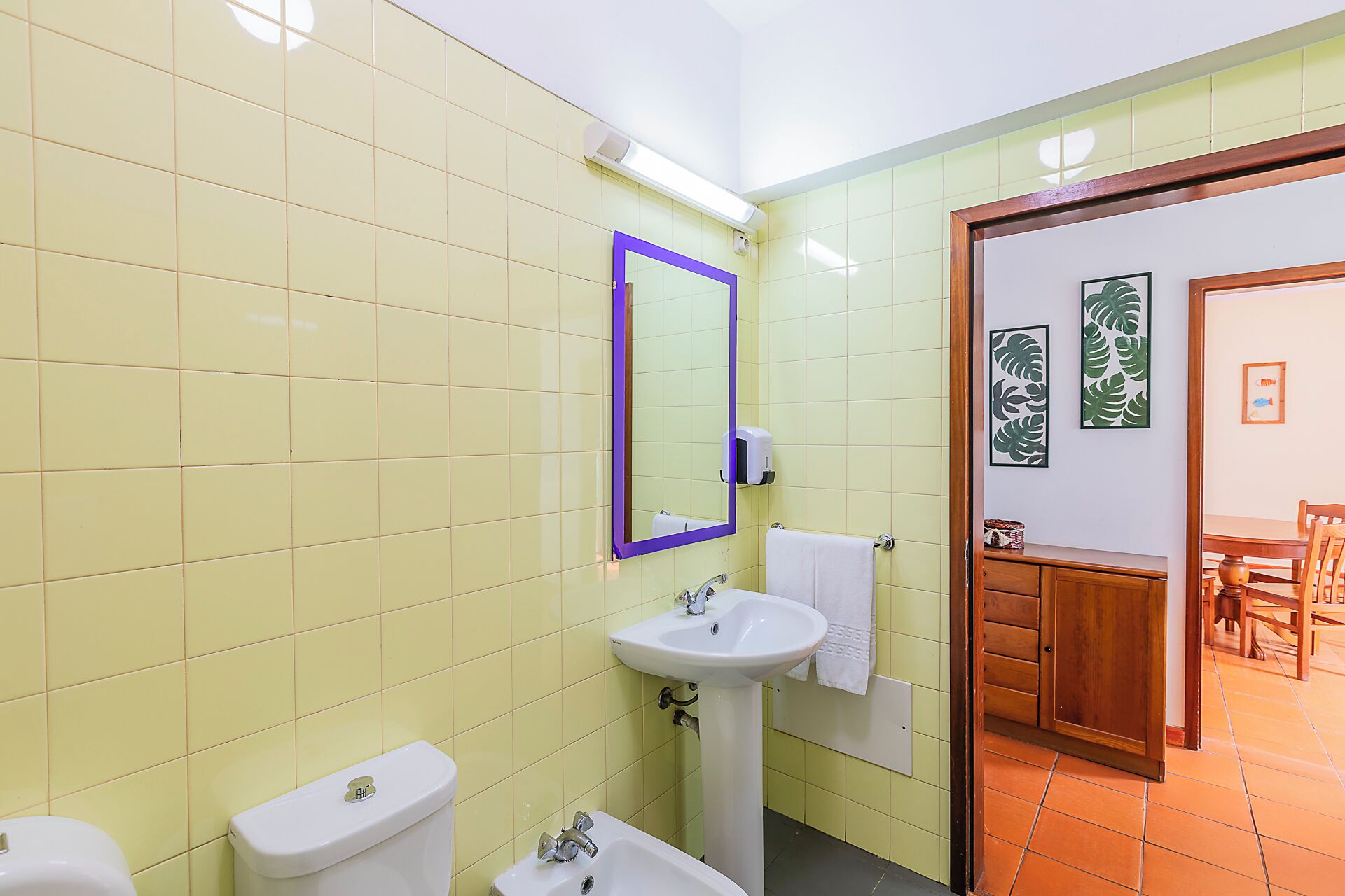 A bathroom with yellow tiles, a white sink, a toilet, and a mirror with a purple frame.