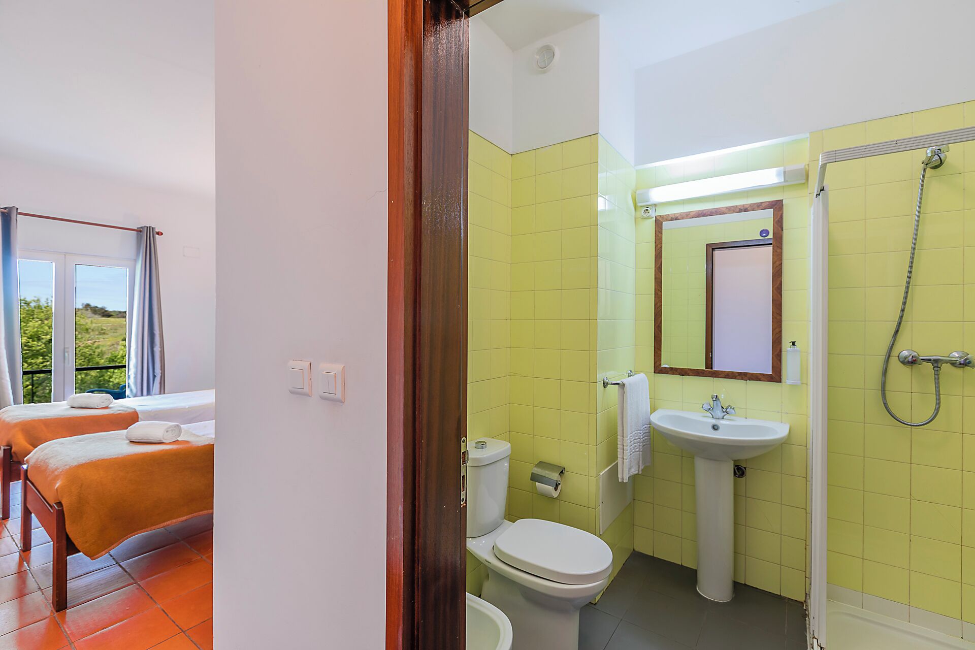 A hotel room with two beds, a bathroom with yellow tiles, a sink, and a shower.