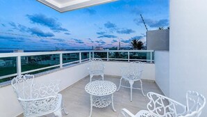 A balcony with white metal chairs and a table, overlooking a body of water and a cityscape. - Aguia Flats (Natal)