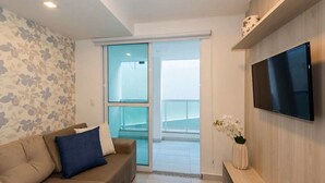 A modern living room with a sofa, a flat-screen TV, a sliding glass door leading to a balcony, and a vase with flowers. - Aguia Flats (Natal)