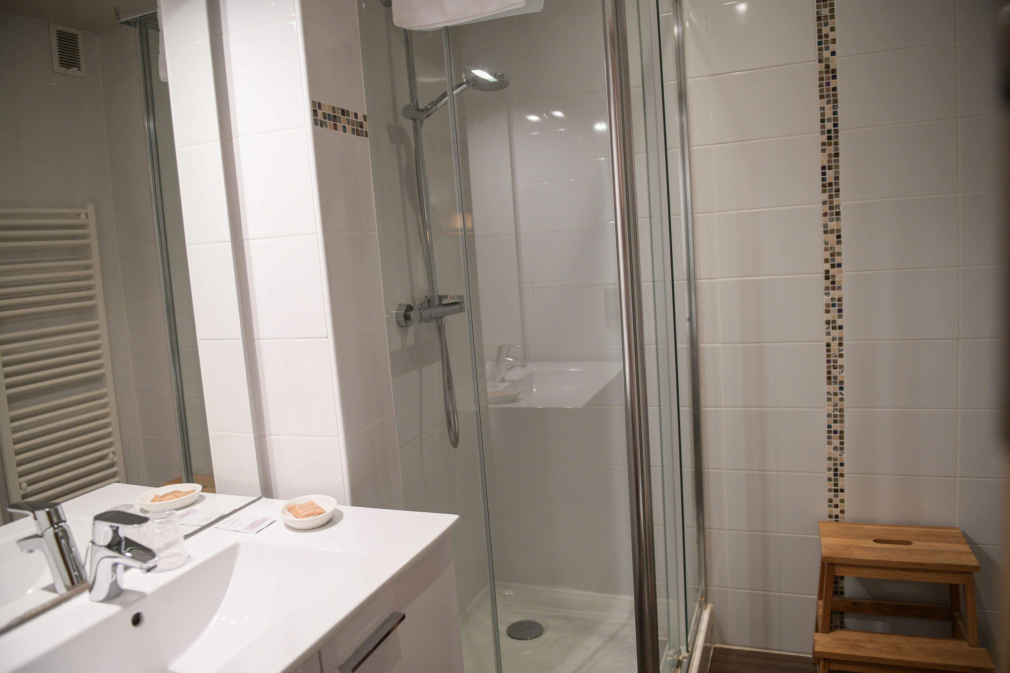 Un bany modern amb dutxa de vidre, un lavabo blanc amb mirall i un petit tamboret de fusta.