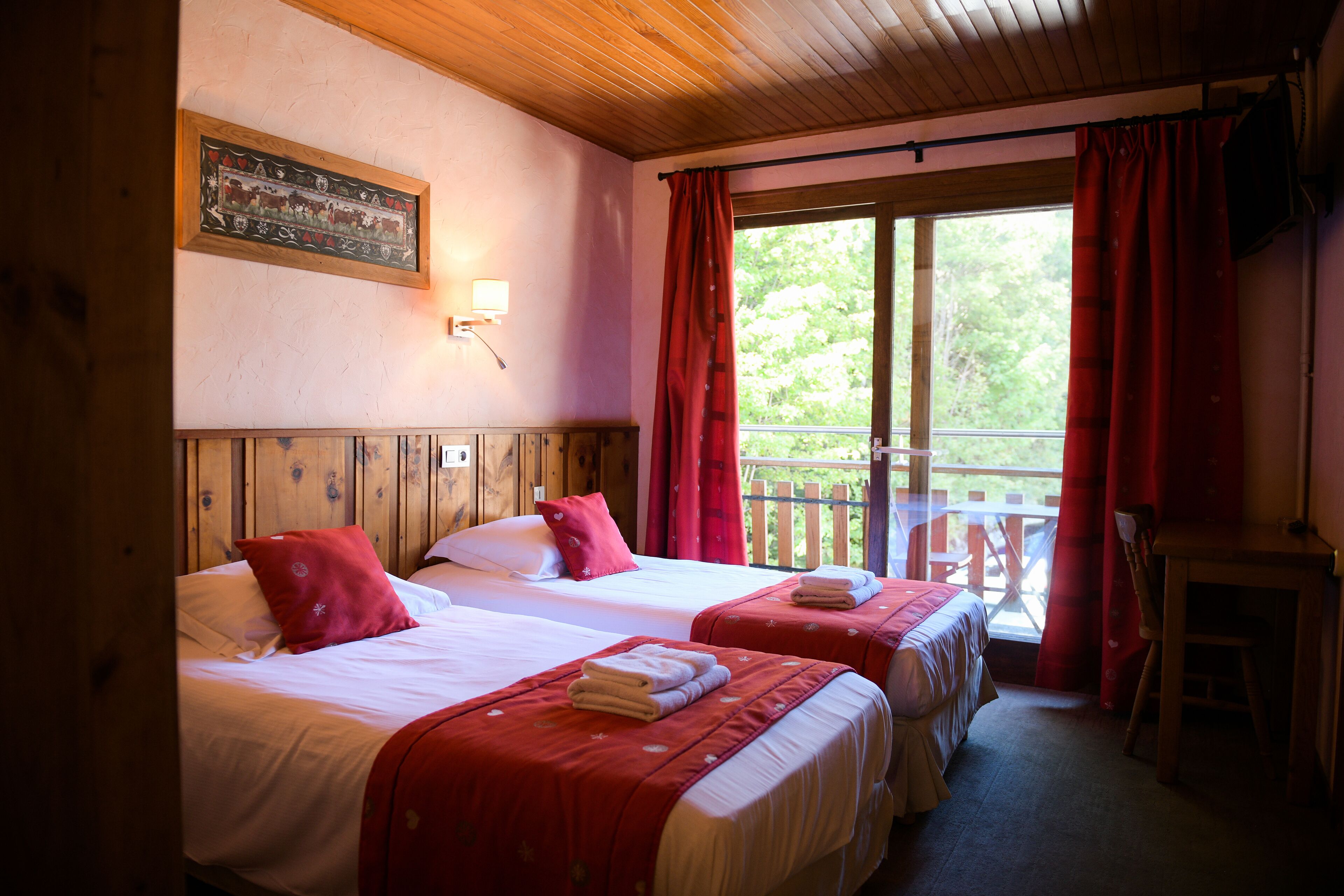 Ein Zimmer mit zwei Betten, Holz-Kopfteilen, einer Holzbalkendecke, einem Fenster mit roten Vorhängen und einem Holztisch.