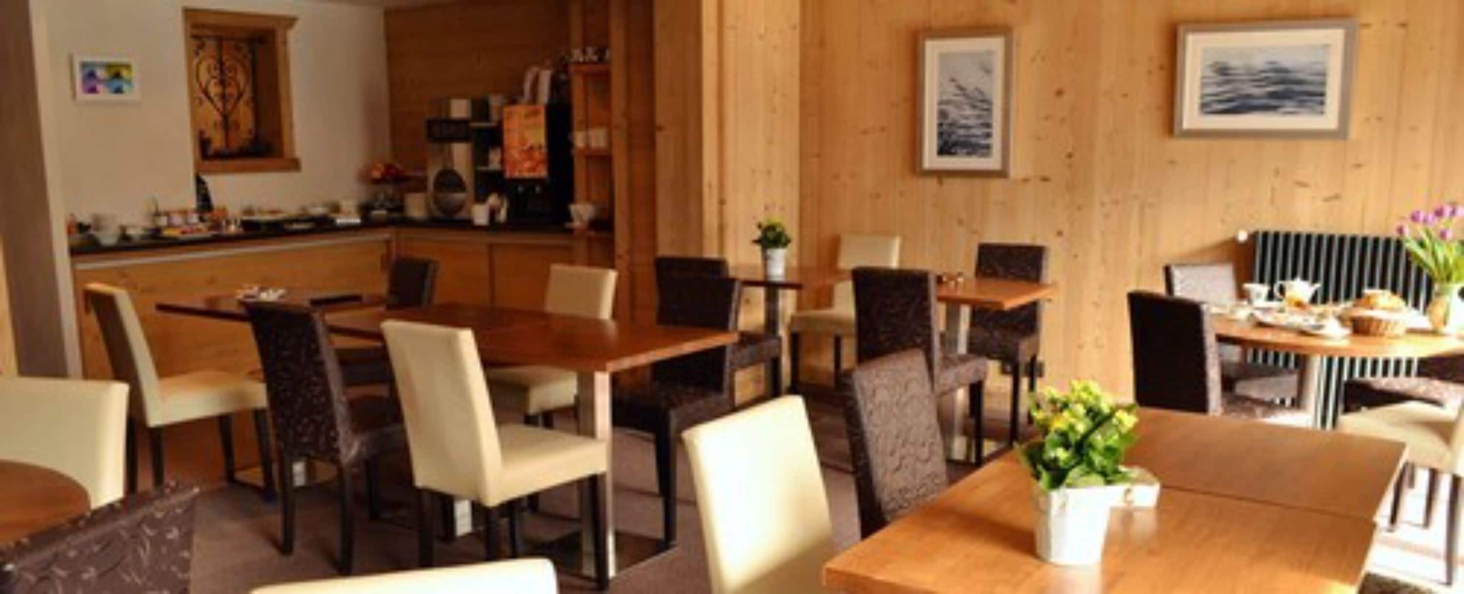 A dining area with wooden tables and chairs, a kitchen in the background, and framed artwork on the walls.