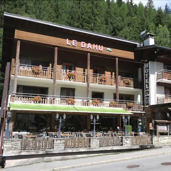 Un hotel llamado "LE DAHU" con fachada de madera, balcones con macetas de flores, asientos al aire libre y un cartel que indica que es un hotel.