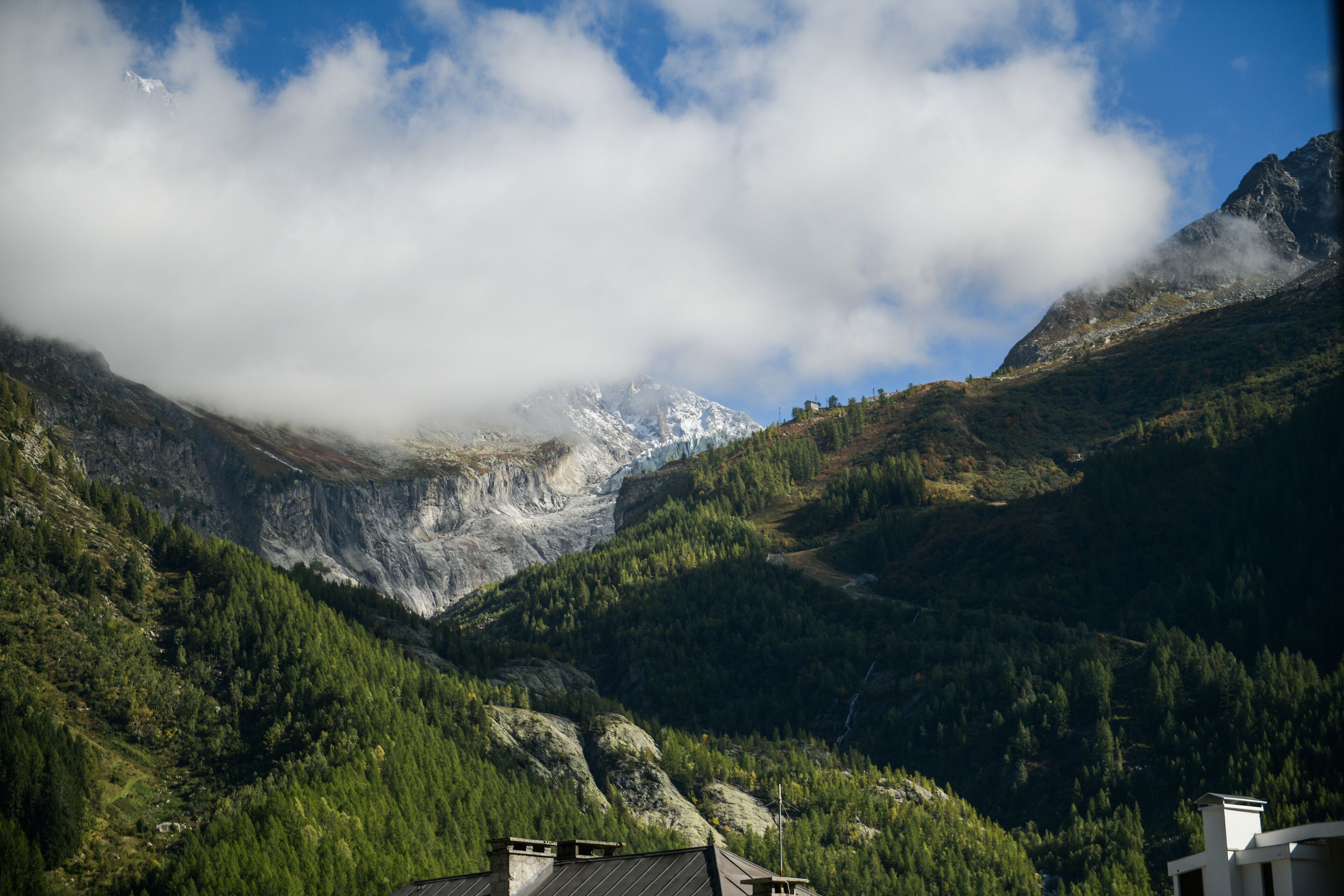 Paisagem montanhosa com vale, uma casa e um pico de montanha parcialmente coberto por nuvens.