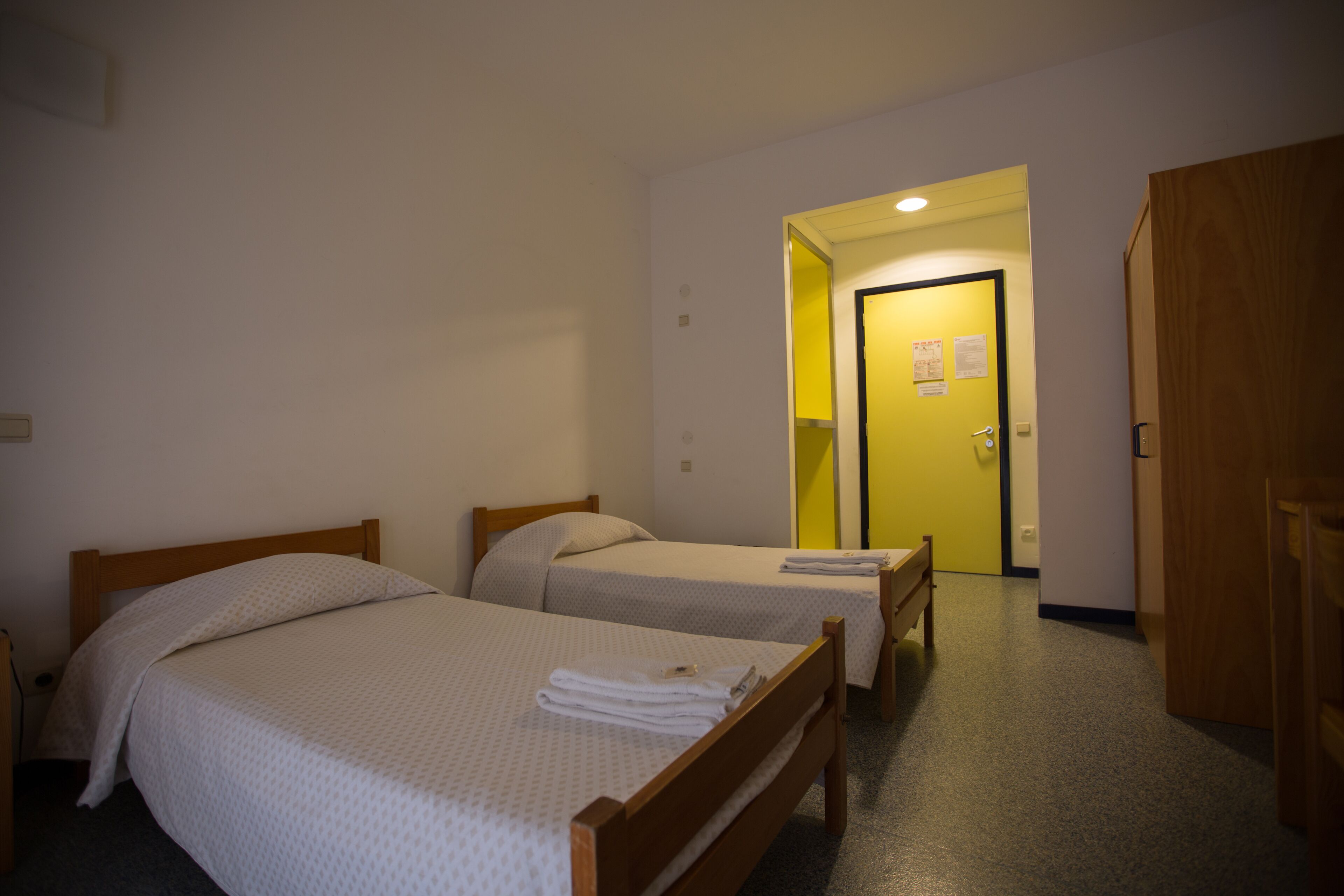 Habitación de hotel con dos camas individuales, un armario de madera y una puerta que da a un pasillo.