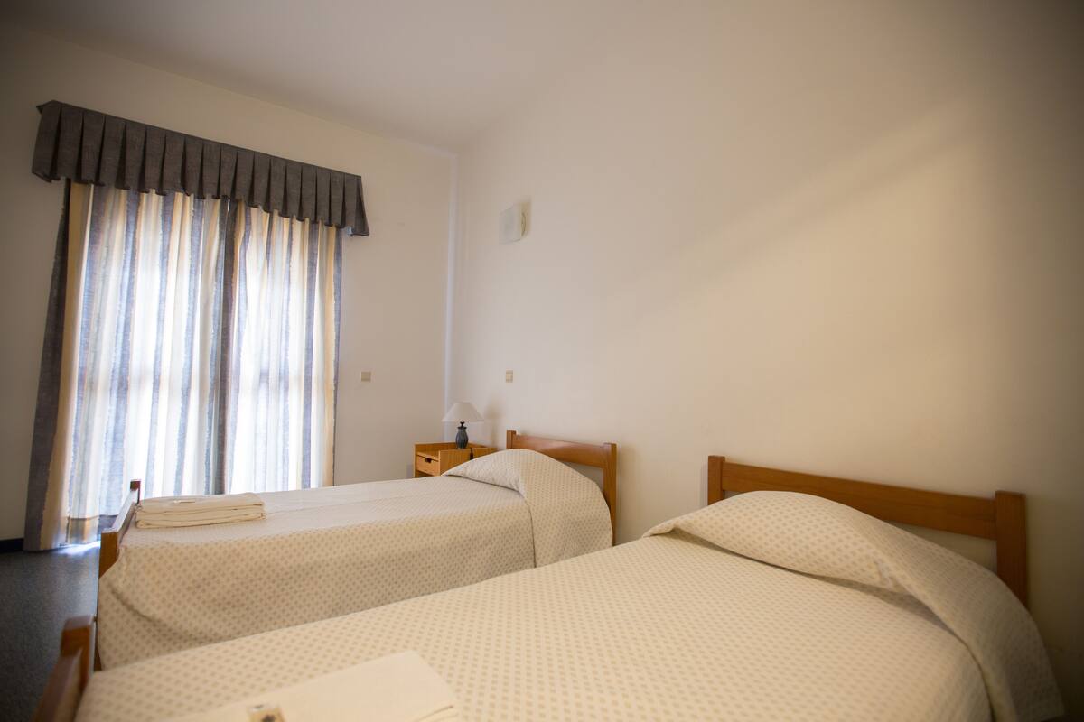 A hotel room with two single beds, a nightstand with a lamp, and a window with curtains.