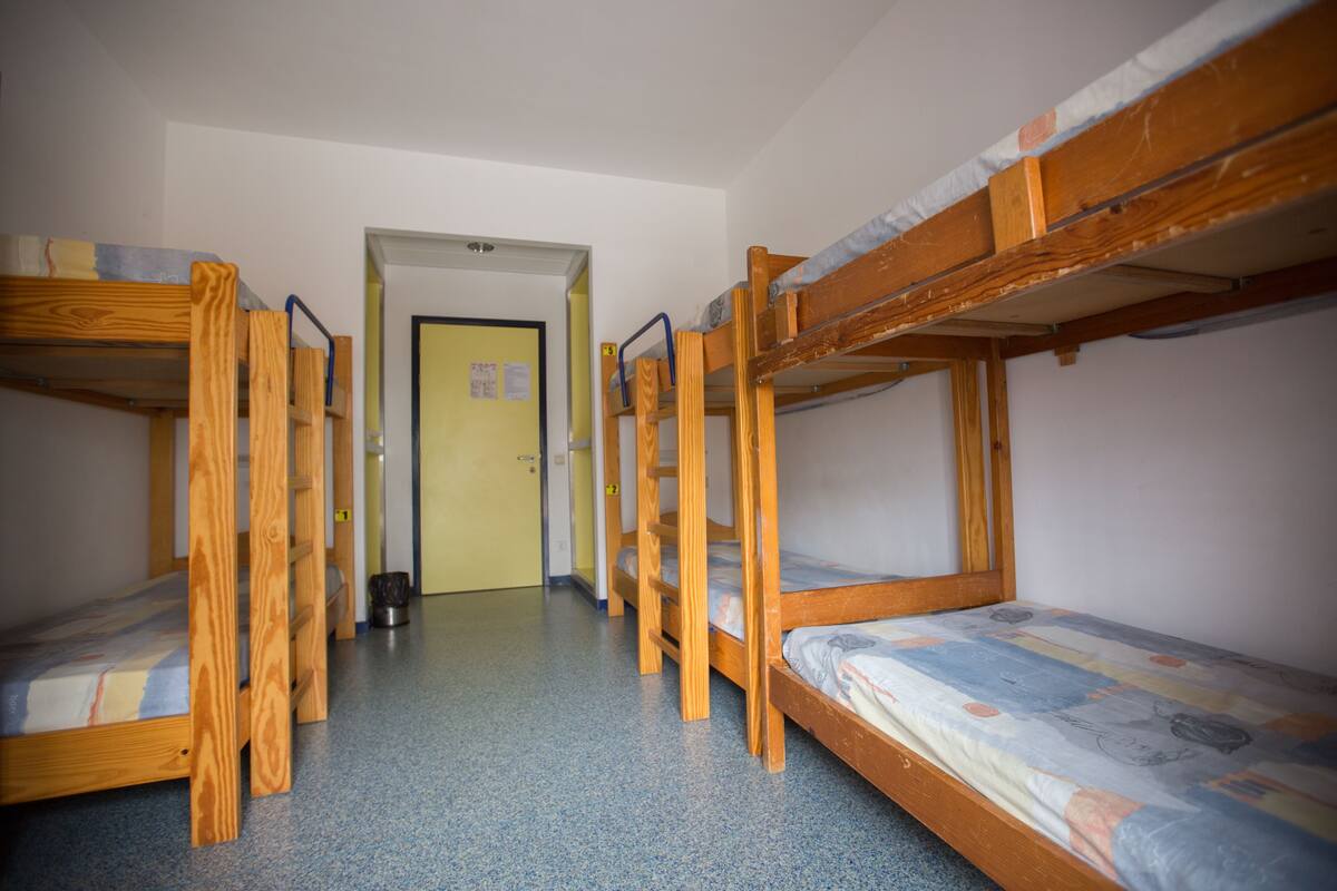 A dormitory with bunk beds arranged in a row, a door in the center, and a trash can near the door.