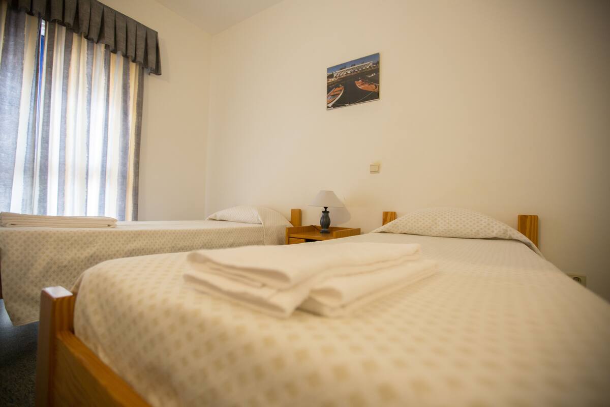 A hotel room with two single beds, a nightstand with a lamp, a window with curtains, and a wall-mounted picture.