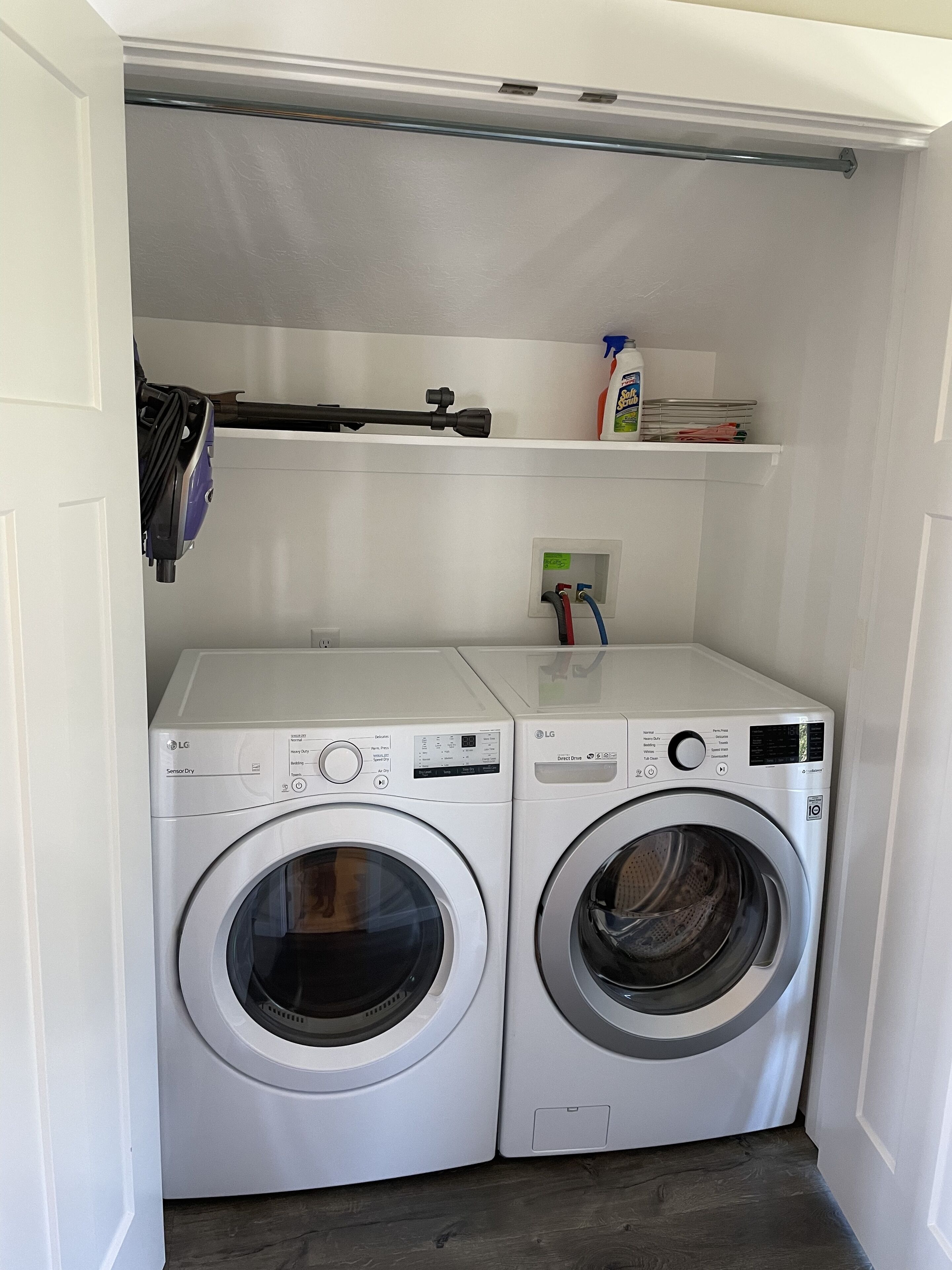 A laundry room with two white LG washing machines, a shelf with cleaning supplies, and a vacuum cleaner.