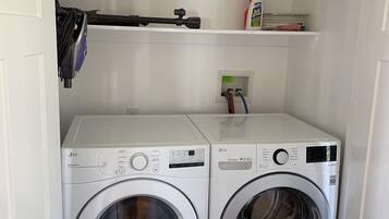 A laundry room with two white LG washing machines, a shelf with cleaning supplies, and a vacuum cleaner.